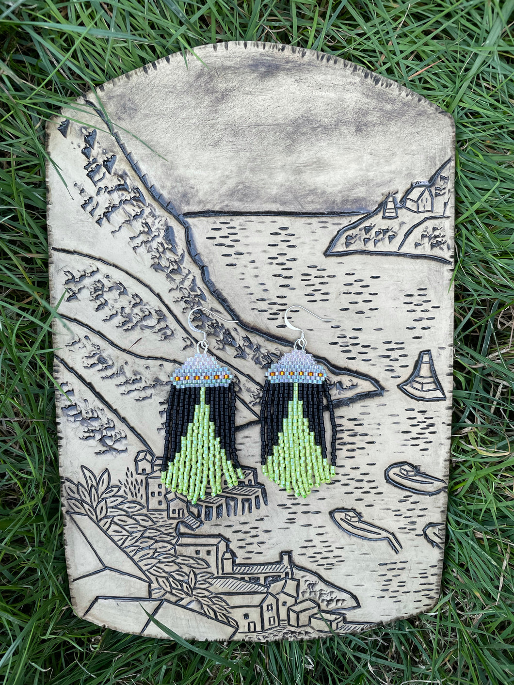 A pair of earrings depicting a UFO with a tractor beam, made from glass beads lays on a ceramic plaque depicting a harbour, which lays on the grass